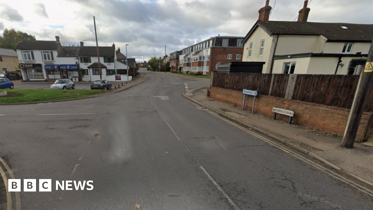 Greenfield Road leading into Flitwick, with houses and small shops on either side of the road and double yellow lines.