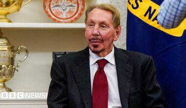 Oracle co-founder Larry Ellison sitting in a chair at the White House wearing a dark grey suit, white dress shirt and deep red neck tie.