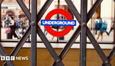 A close-up shot of a London Underground roundel, seen through the diamond patterns of a closed metal concertina gate. In the blurred background, people walk past.