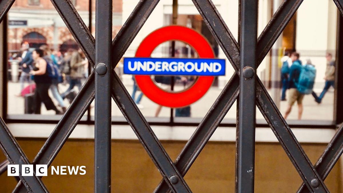 A close-up shot of a London Underground roundel, seen through the diamond patterns of a closed metal concertina gate. In the blurred background, people walk past.