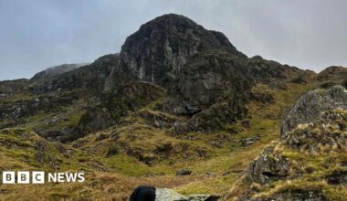 Priest's Hole at Dove Crag in the Lake District. It is a mossy, black rock face that has broken, grassy rocky ground leading up to it.