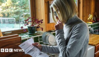 Woman looks at a bill in her kitchen with plates on the draining board behind her.