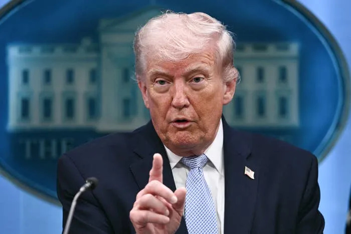 A person in a suit gestures during a speech at a podium, with a blurred White House emblem in the background