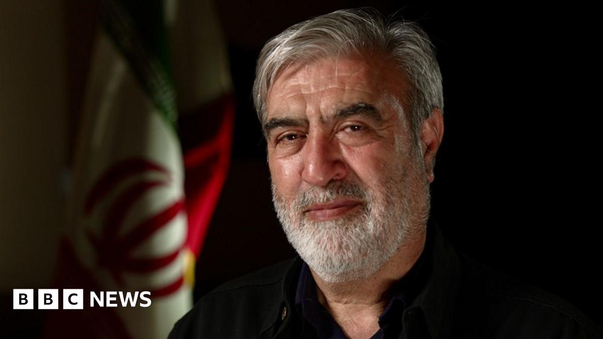 A man with a grey beard and hair looks into the camera. He is wearing a black shirt and behind him is the Iranian flag.
