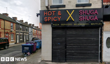 A shop has a sign saying 'Hot and Spicy', 'X' and 'Shuga Shuga' with 'pizzas - burgers - milkshake - waffles' underneath. Black shutters are down across the shop front. Industrial waste bins can be seen in a side street.