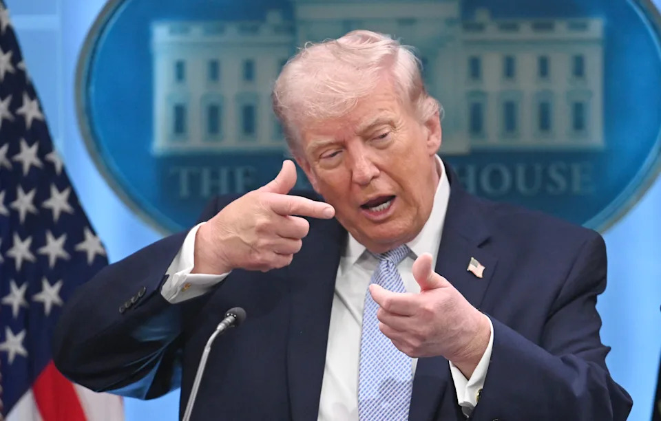 Man gestures during a press conference at a podium with the presidential seal. Background shows American flags and a White House emblem