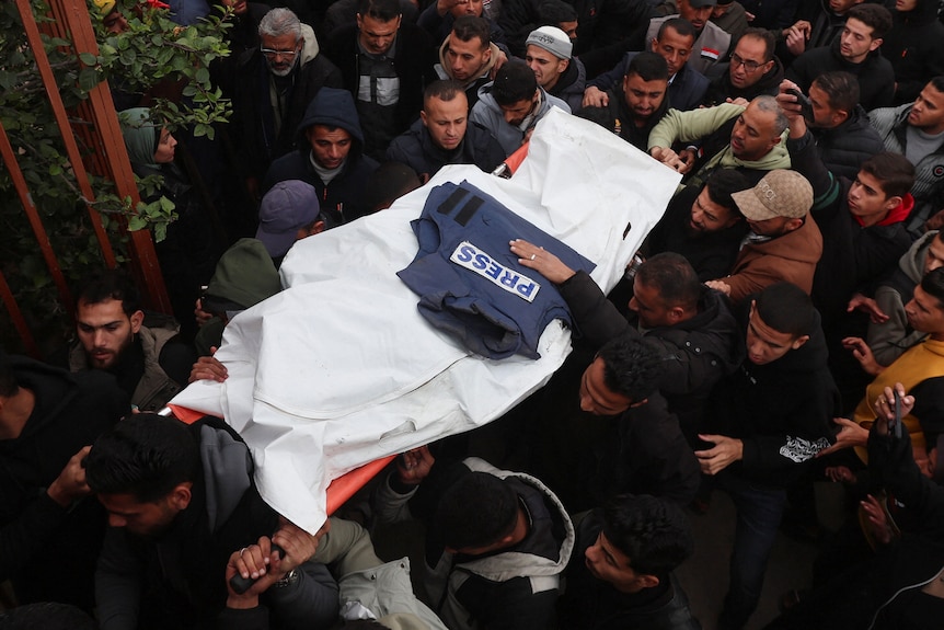 A crowd of people carry a stretcher covered by a while cloth with a press flak-jacket on top.