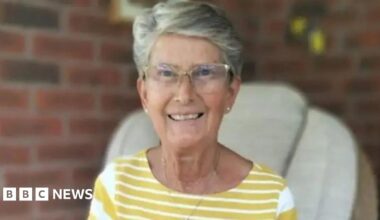 A woman with short grey hair and clear glasses is smiling at the camera and sitting in a chair. She is wearing a yellow and white-striped top and a necklace with white-stud earrings. The background is blurred but there is a brick wall behind her.