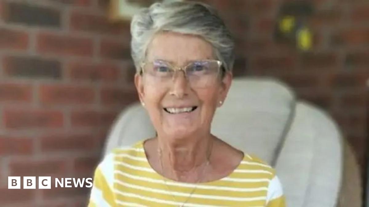 A woman with short grey hair and clear glasses is smiling at the camera and sitting in a chair. She is wearing a yellow and white-striped top and a necklace with white-stud earrings. The background is blurred but there is a brick wall behind her.