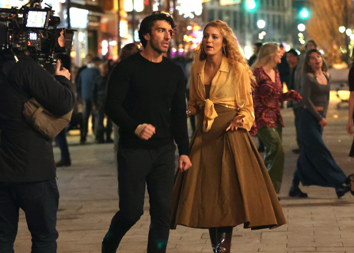 Blake Lively filming a dramatic scene at night, wearing a brown skirt and yellow blouse, surrounded by people and camera crew.