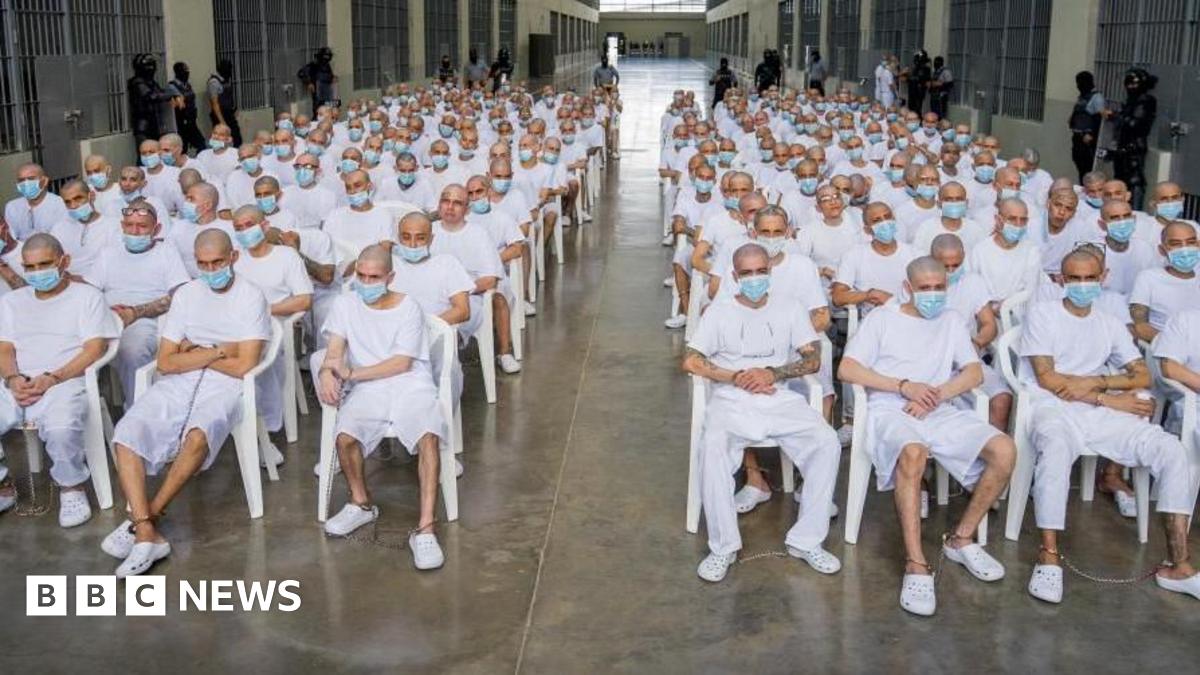 A group of hundreds of men with shaved heads, wearing white t-shirts and shorts or trousers and blue surgical masks, are seen sitting on white plastic chairs in an open space at a confinement centre in El Salvador