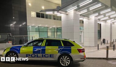 A police car parked outside the Royal Victoria Hospital Emergency Department. The building is white. The police car is yellow and blue and says POLICE along the side of it.
