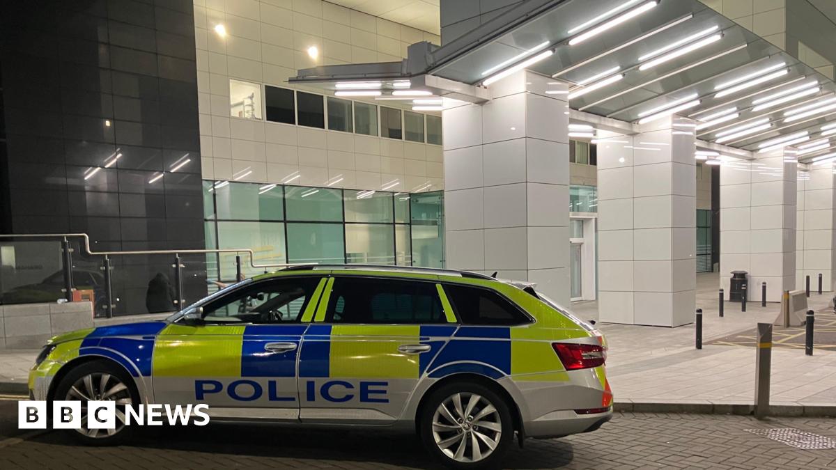 A police car parked outside the Royal Victoria Hospital Emergency Department. The building is white. The police car is yellow and blue and says POLICE along the side of it.