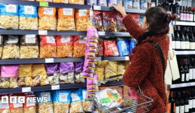 People walk across a bridge over the Thames in a blur as a red double decker bus drives past, with the sun low in the sky casting long shadows.