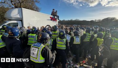 White truck with two people sat on the roof - dozens of police officers in riot gear are standing in a group with a large crown seen beyond in a field.