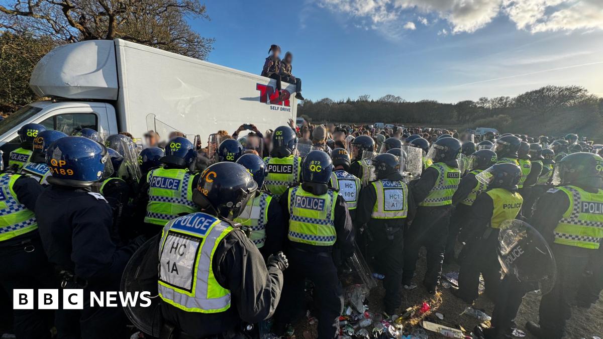 White truck with two people sat on the roof - dozens of police officers in riot gear are standing in a group with a large crown seen beyond in a field.