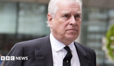 Andrew Mountbatten-Windsor, with short white hair in a black suit and tie, walks past a glass building