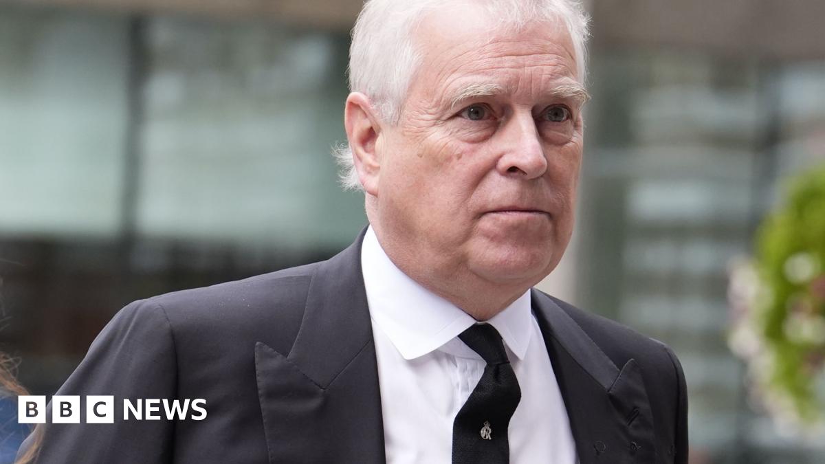 Andrew Mountbatten-Windsor, with short white hair in a black suit and tie, walks past a glass building