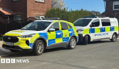 A police car parked in front of a police van. The car is a large, white Ford with bright yellow and blue squares covering most of the car. The van is mostly white with the same pattern. They are parked half on the curb in front of a red brick semi-detached house with a brown fence.