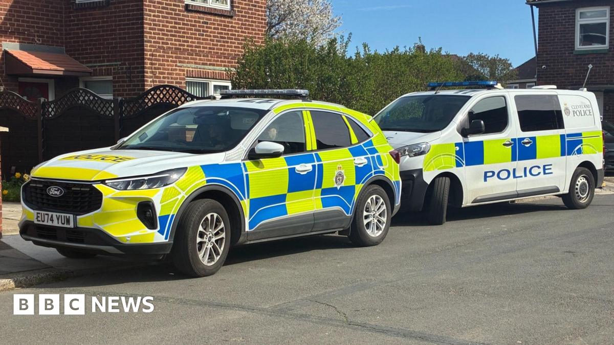 A police car parked in front of a police van. The car is a large, white Ford with bright yellow and blue squares covering most of the car. The van is mostly white with the same pattern. They are parked half on the curb in front of a red brick semi-detached house with a brown fence.