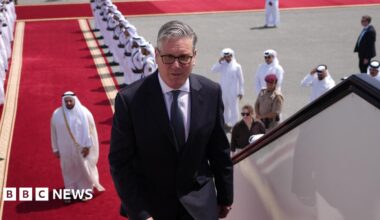 Keir Starmer boarding a plane at the airport in Doha, Qatar. There is a red carpet, lined with men, leading up to the plane