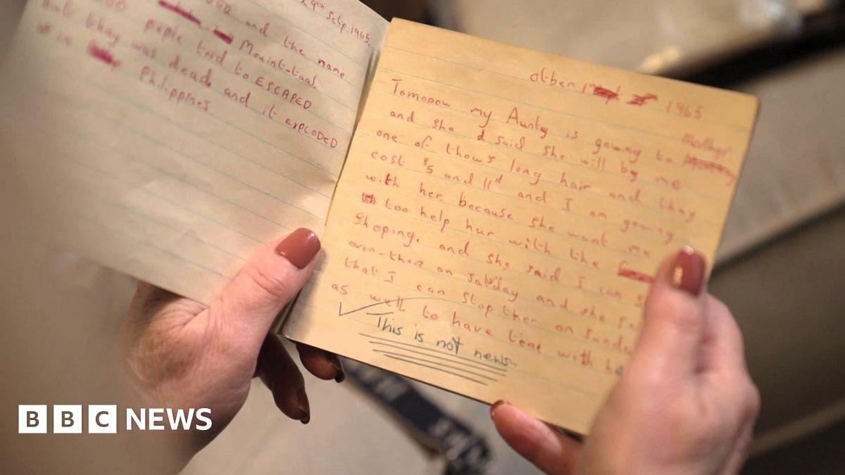 A close up of a school exercise book from the 1960s, the pages are yellowed. One page is filled with text handwritten in red ink, which details a trip to the shops. At the bottom of the page, underlined, a teacher has written "This is not news".