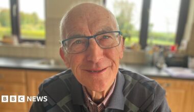 Alphie Lonergan sits at his kitchen table with his sons Ronan and Donal. Donal, in the middle, is wearing a navy quarter-zip top and has receding dark hair. Ronan is wearing a striped top and is bald on top with dark hair at the sides. The kitchen table has a grey/green tablecloth with white polka dots on it and there is a plate of biscuits in the middle. A mug is in front of each of the three men.