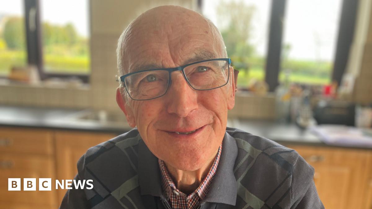 Alphie Lonergan sits at his kitchen table with his sons Ronan and Donal. Donal, in the middle, is wearing a navy quarter-zip top and has receding dark hair. Ronan is wearing a striped top and is bald on top with dark hair at the sides. The kitchen table has a grey/green tablecloth with white polka dots on it and there is a plate of biscuits in the middle. A mug is in front of each of the three men.