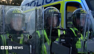 Police officers wearing helmets and high‑visibility vests stand close together holding clear riot shields, with a marked police van parked behind them.