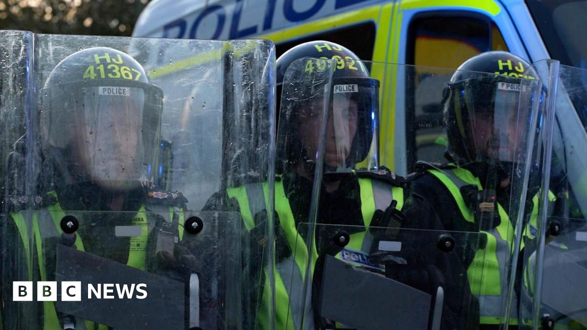 Police officers wearing helmets and high‑visibility vests stand close together holding clear riot shields, with a marked police van parked behind them.