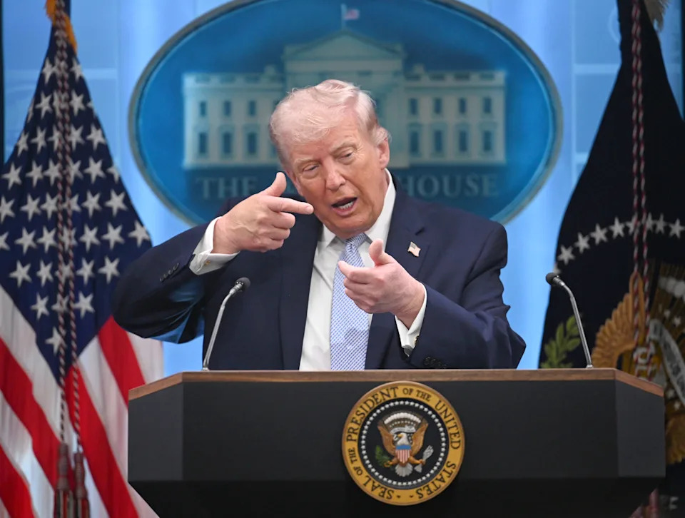 A person at a press briefing makes a gesture with their hand, standing at a podium with the White House emblem in the background