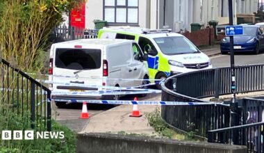 Two police vans are parked close to each other near a pavement with a dark fence running alongside it. There are cones and several lines of police tape running across the street.