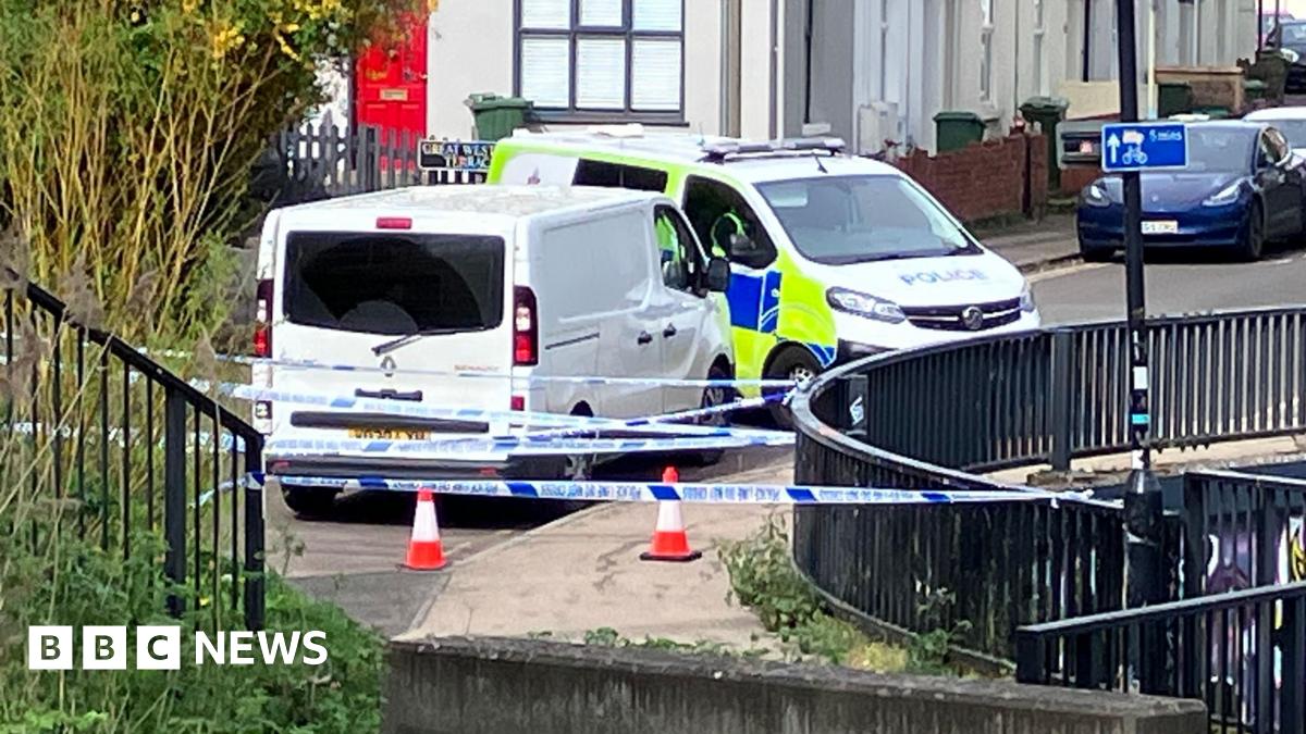 Two police vans are parked close to each other near a pavement with a dark fence running alongside it. There are cones and several lines of police tape running across the street.