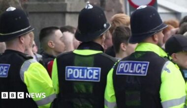 A picture of football fans crowded together in a group. They are stood behind a line of police officers. They are wearing green hi-vis vests on top of their clothes.
