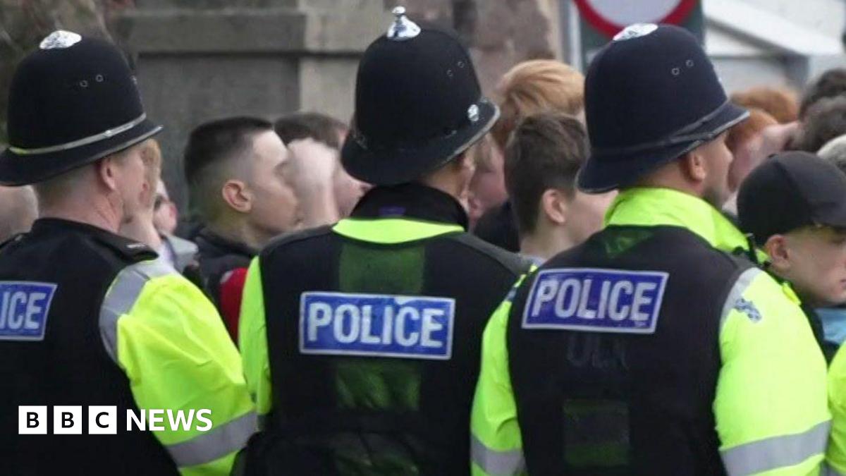 A picture of football fans crowded together in a group. They are stood behind a line of police officers. They are wearing green hi-vis vests on top of their clothes.