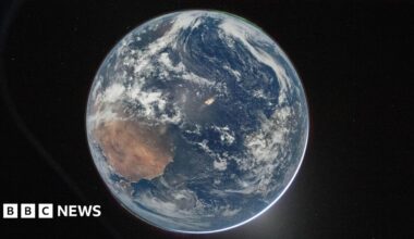 A picture of the Earth from space, which centres the planet against the dark background of space. It's a round blue planet. Clouds can be seen and a thin green aurora at the top.