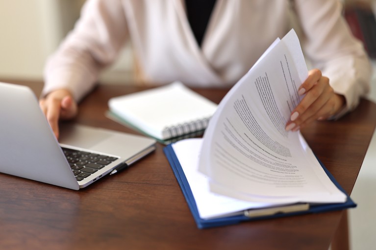 An unidentifiable woman is working at a desk with a laptop and notepad while reviewing a paper.