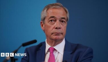 Nigel Farage is sat down in front of a microphone and a blue backdrop. He has short grey hair and is wearing a dark blue suit, white shirt and a pink tie.