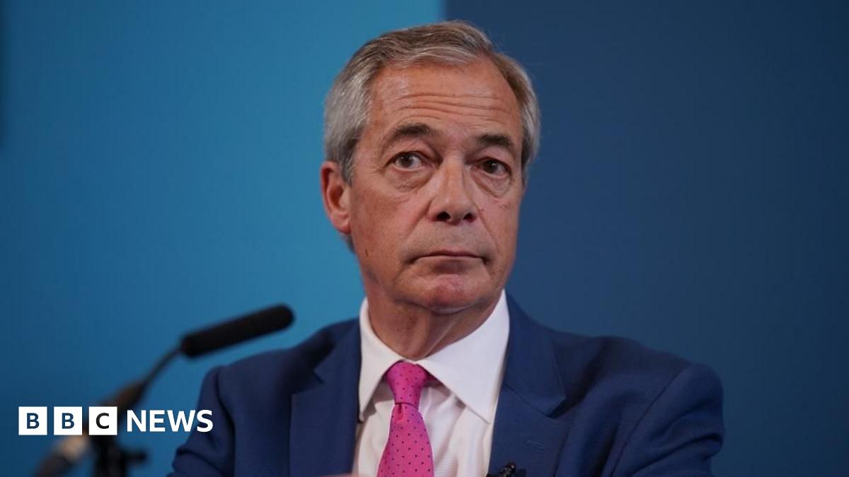 Nigel Farage is sat down in front of a microphone and a blue backdrop. He has short grey hair and is wearing a dark blue suit, white shirt and a pink tie.