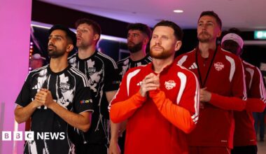 Three members from the Sidemen on the left dressed in a black jersey and three from the YouTubers Allstars dressed in red, standing in the tunnel with their hands together.