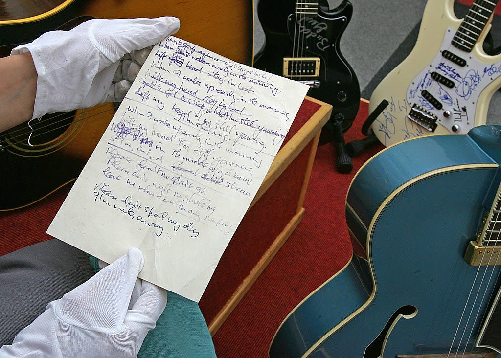 A person wearing gloves holds a handwritten note among several signed guitars, suggesting a music memorabilia collection