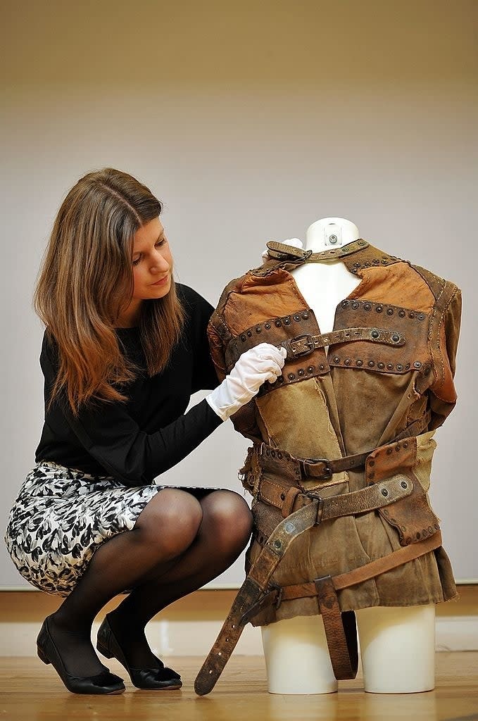 Woman examines a vintage straitjacket on a mannequin in a museum setting. She wears a black top, patterned skirt, and white gloves