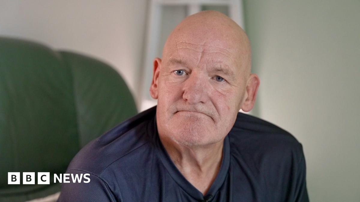 Patrick Moore sitting on a green chair looking at the camera wearing a navy blue top. He has a bald head and is looking glum at the camera.