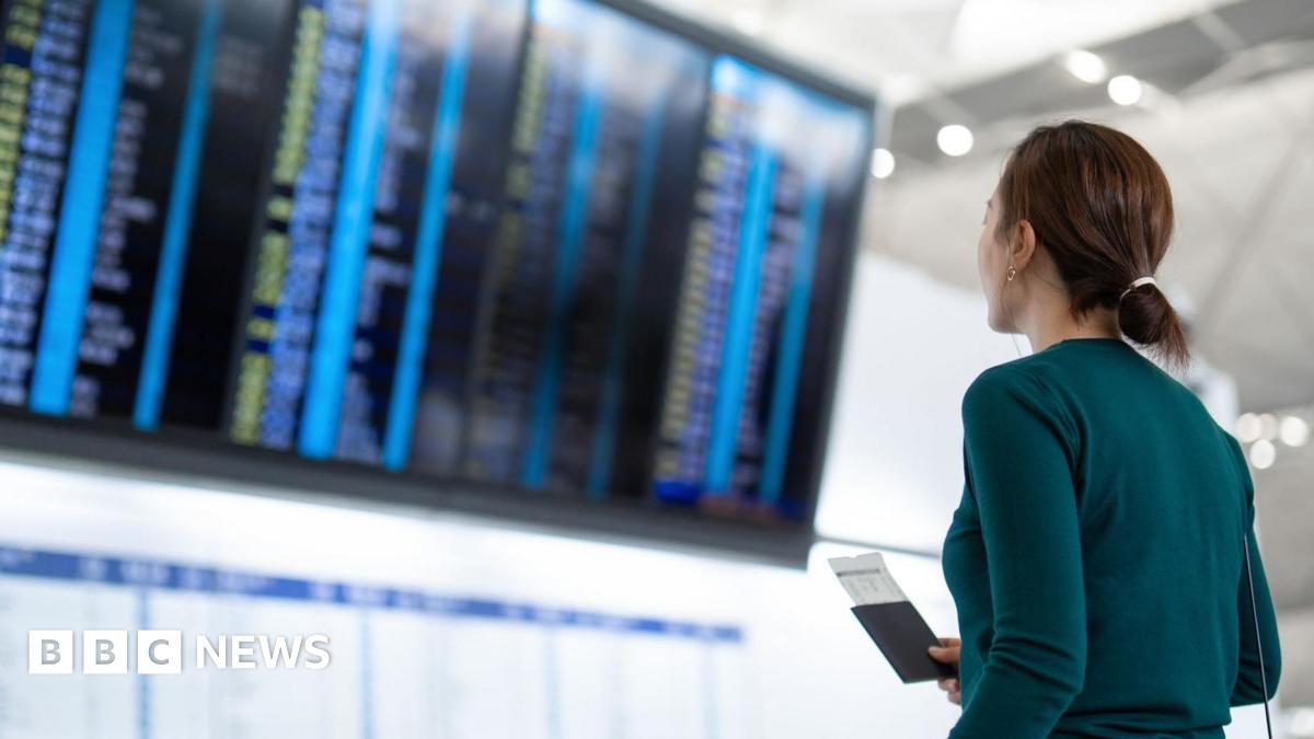 A woman wearing a dark turquoise top is seen from behind, standing looking at an airport departures board. She is holding her passport and boarding pass