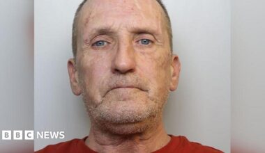 Stephen looks at the camera with a blank expression. He has short grey hair, a stubbly beard, and wears a red t-shirt.