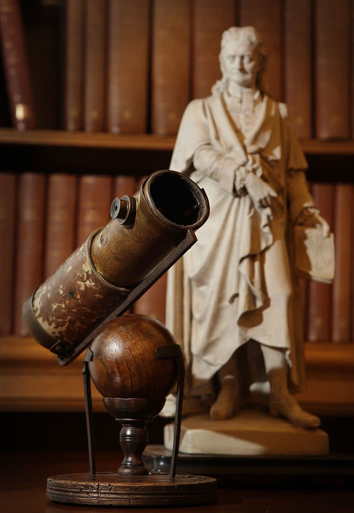 A telescope is displayed in front of a statue of a historical figure, set against a background of bookshelves