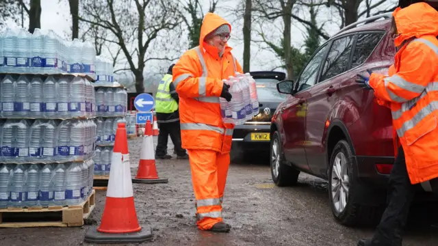 A woman in a orange high visibility suit. She is carrying a pack of large water bottles.