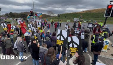 A group of protestors seen mostly from behind as they face a grassy bank. They stand in the road between traffic lights holding black and white CND signs saying Kick out Trump's Nukes and No War on Iran.