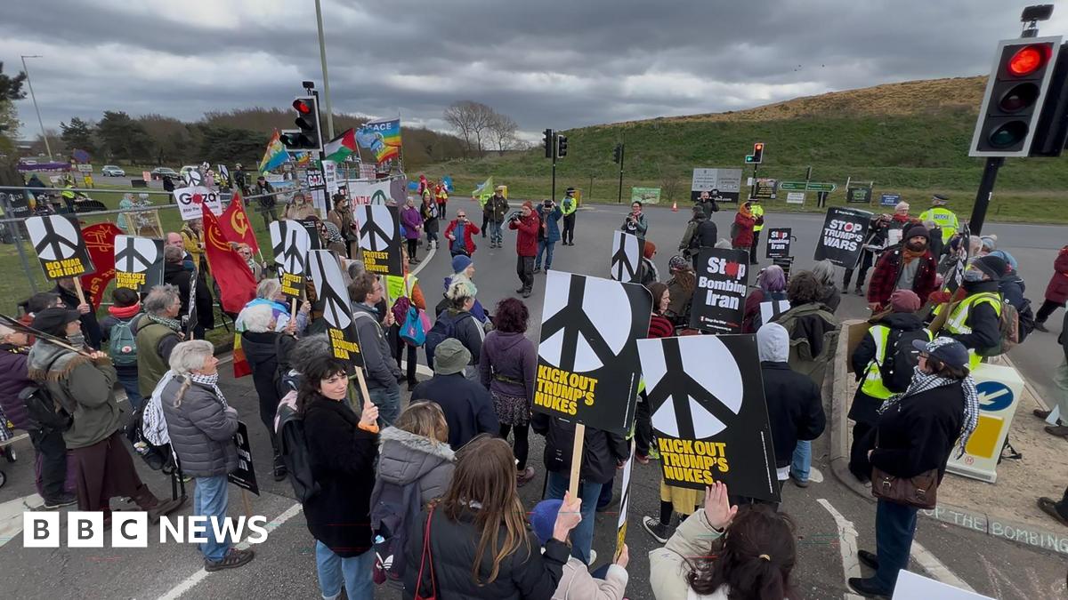 A group of protestors seen mostly from behind as they face a grassy bank. They stand in the road between traffic lights holding black and white CND signs saying Kick out Trump's Nukes and No War on Iran.