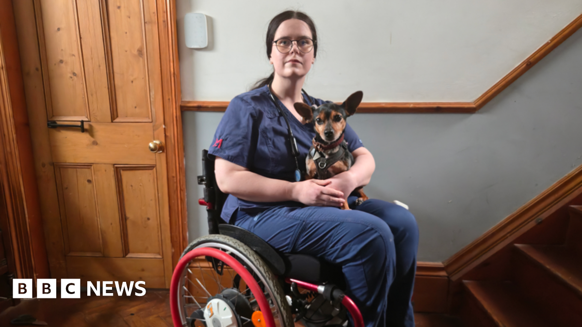 A woman with tied-back brown hair is wearing a dark blue surgical gown and sitting in a red wheelchair. She has a small terrier-type dog on her lap. They are in a corridor with wooden flooring and stairs.